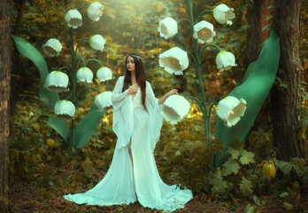 Beautiful fantasy woman princess elf in long white dress walks in fairy forest with large flowers...