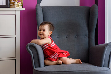 Niña pequeña simpática y sonriente con vestido rojo con lineas blancas sentada en sillón gris