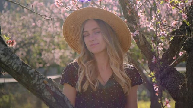 Soft and romantic shot of young beautiful woman in sun flare sunshine. Authentic real millennial woman look at camera, wear straw hat and dress in flower blossom trees. Spring summertime concept
