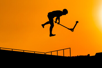 Unrecognizable teenage boy silhouette showing high jump tricks on scooter against orange sunset sky at skatepark. Sport, extreme, youth, urban culture, freestyle, outdoor activity concept