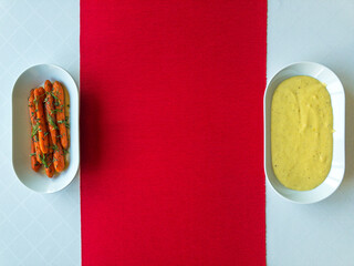 Glazed carrots with dill and thyme, and mashed potatoes with black pepper, white pepper and nutmeg on white tablecloth and red table runner, top view