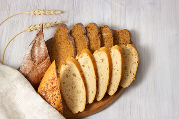 Slices of fresh homemade baguettes with whole grain and seeds on wooden board with ears of wheat. White wooden background. Top view with copy space.