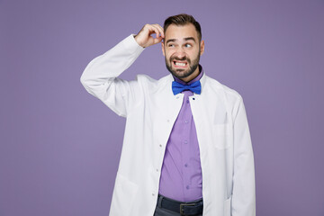 Preoccupied puzzled young bearded doctor man wearing white medical gown put hand on head looking aside isolated on violet wall background studio portrait. Healthcare personnel health medicine concept.