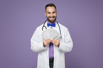 Smiling young bearded doctor man in medical gown stethoscope hold fan of cash money in dollar banknotes isolated on violet background studio portrait. Healthcare personnel health medicine concept.