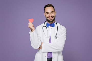 Smiling young bearded doctor man wearing white medical gown stethoscope hold red little heart isolated on violet colour wall background studio portrait. Healthcare personnel health medicine concept.