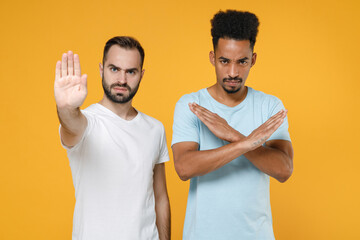 Displeased young two friends european african american men 20s in white blue casual t-shirts showing stop gesture with palm crossed hands isolated on bright yellow colour background studio portrait.