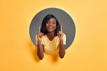 Close-up view portrait of young cheerful woman pointing two fingers up isolated in a round hole in bright yellow background. Copy space.