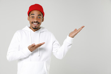 Excited cheerful smiling young african american man 20s wearing basic casual streetwear hoodie pointing hands aside on mock up copy space isolated on white colour wall background, studio portrait.