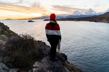 mujer chica al atardecer asomada a precipicio en el mar con gorro rojo y jersey de lana