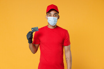 Fototapeta na wymiar Delivery employee african man in red cap blank print t-shirt face mask gloves work courier service on quarantine covid-19 concept hold mockup of credit bank card isolated on yellow background studio.