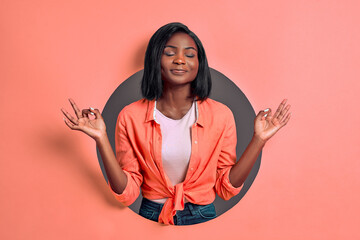 Portrait of beautiful calm young  female practicing yoga indoors, meditating, holding hands in mudra gesture isolated in a grey circle on a coral background. Soul and mind zen balance concept.