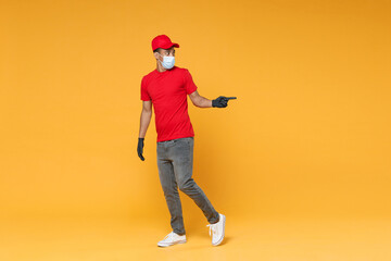 Fototapeta premium Full length delivery employee african man in red cap t-shirt face mask gloves uniform work courier dealer service on quarantine coronavirus covid-19 virus concept isolated on yellow background studio.