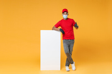 Full length delivery employee african man in red cap t-shirt face mask gloves work courier dealer service hold big white empty blank billboard for promo copy space isolated on yellow background studio