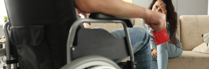 Sad young woman sits on couch opposite man in wheelchair with condom in hand. Sexual impotence of...