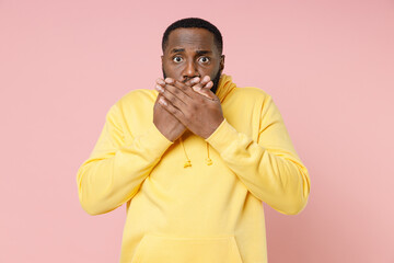 Shocked worried young african american man 20s wearing casual basic yellow streetwear hoodie standing covering mouth with hands looking camera isolated on pastel pink color background studio portrait.