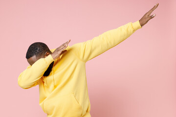 Young african american man 20s in casual yellow streetwear hoodie doing dab hip hop dance hands gesture, youth sign hiding and covering face isolated on pastel pink color background studio portrait.