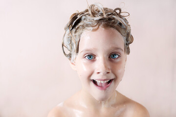 a little blonde girl with a soapy head smiles in the bathroom on a pink background