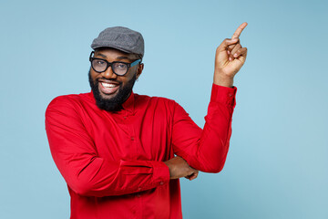 Naklejka premium Smiling cheerful young bearded african american man 20s wearing casual red shirt eyeglasses cap pointing index finger up on mock up copy space isolated on pastel blue color background studio portrait.