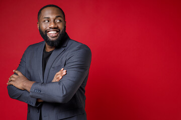 Side view of smiling attractive young african american business man 20s in classic jacket suit standing holding hands crossed looking aside isolated on bright red color background studio portrait.