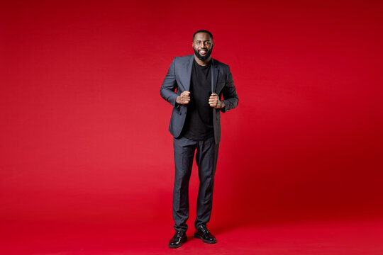 Full Length Of Smiling Attractive Confident Young African American Business Man 20s Wearing Classic Jacket Suit Standing Looking Camera Isolated On Bright Red Color Wall Background Studio Portrait.