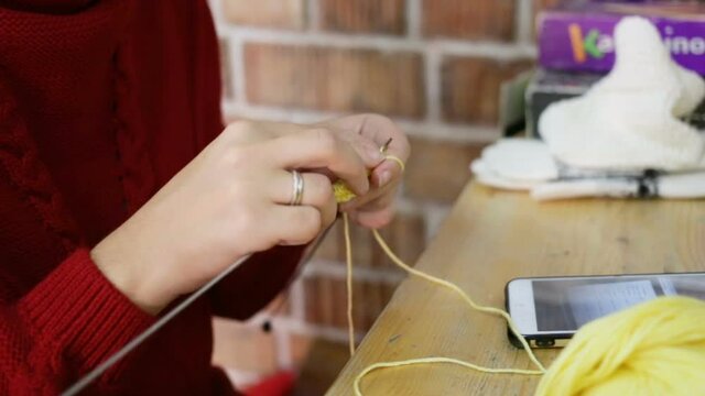 A woman pulls a ball of thread, she knits a yellow baby bootie. On the table are: a Cup of coffee, a smartphone, cookies, a skein of wool thread. Close-up, real-time, contains people in the room