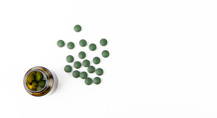 Green spirulina tablets scattered on a white background from a glass jar.