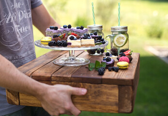 Unrecognizable man carrying outdoors selection of colorful and delicious cake desserts on tray.