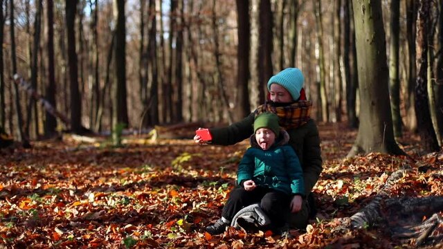 Young happy mother with little cute boy using smart phone mobile sitting in autumn forest enjoying nature beauty on sunny day. Blogging motherhood. Video call. Kids selfie. True feelings.