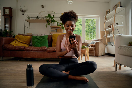 Portrait of smiling young african woman using smartphone after routine wokout with bottle besides at home