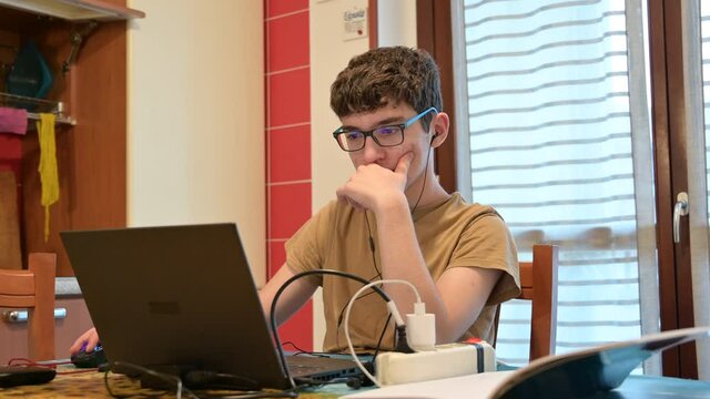 Secene of life at home, a caucasian boy with curly hair and glasses is following the school lesson on the laptop next to a notebook. He set up his study station on the kitchen table.