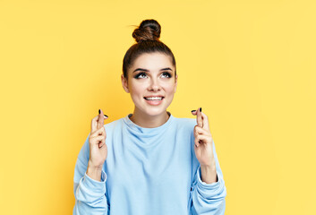 Young beautiful woman crossing her fingers and wishing for good luck over yellow background.
