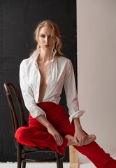 A beautiful blonde curly girl wearing a red pants and a blouse, unbuttoned on her braless big breasts, poses sitting on the chair in the studio. Advertising, fashionable, commercial, design