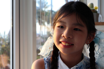 an asian smiling long hair young kid girl is sitting on the wooden chair with background of big window , trees and sky in the cafe coffee shop in the evening