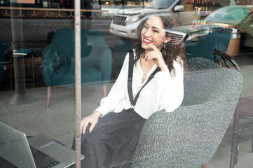 Beautiful curl woman making notes and working in cafe. She is sitting at table with laptop. Freelance concept. View from street through window glass.