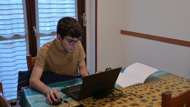 Secene of life at home, a caucasian boy with curly hair and glasses is following the school lesson on the laptop next to a notebook. He set up his study station on the kitchen table.