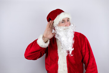 Man dressed as Santa Claus standing over isolated white background surprised with hand over ear listening an hearing to rumor or gossip