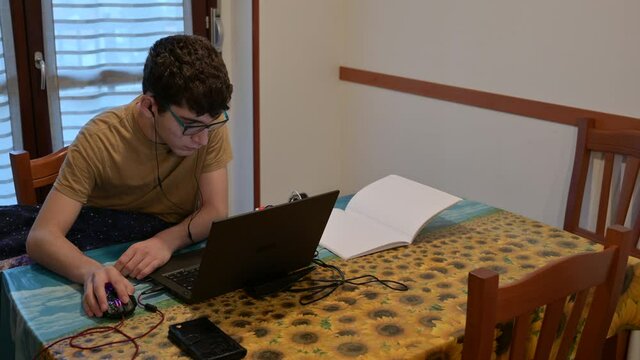 Secene of life at home, a caucasian boy with curly hair and glasses is following the school lesson on the laptop next to a notebook. He set up his study station on the kitchen table.