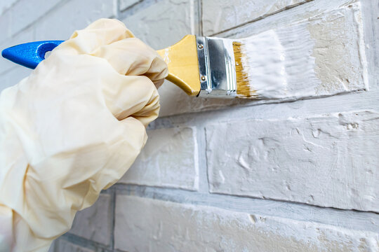 Worker's hand in light rubber gloves paints with a brush a brick wall of white paint, repair concept. Interior renovation, working tools close-up