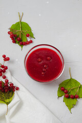 Viburnum jam in glass bowl on the light grey background decorated with Viburnum branches, leaves, and white towel. Autumn composition top view