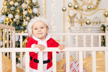 a child in a Santa costume stands in a crib at home near a Christmas tree with a Golden decor