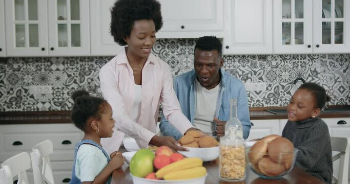 Beautiful cheerful young black-skinned mother putting on the table tasty biscuits for breakfast while her joyful family sitting around the kitchen table and waiting fo them,close up,slow motion