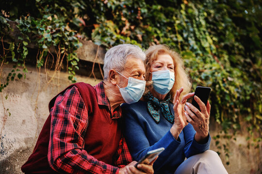 Cute senior couple in love wearing protective surgical masks and looking at smart phone. Seniors using new technologies concept.