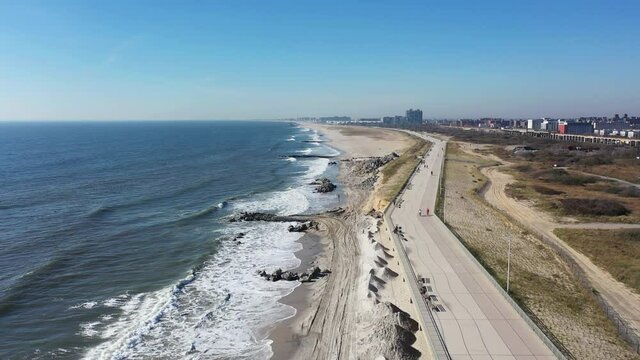An aerial view over the beach in Far Rockaway, NY. It is a sunny day with blue skies. The camera dolly in between the shore & the boardwalk. It is a peaceful scene with only a few people in view.