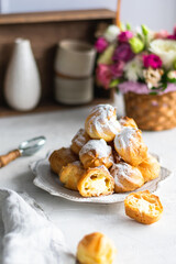 Homemade profiteroles with cream in a gray plate on a white table. Basket with fresh flowers