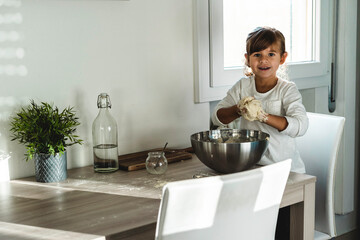 Fototapeta na wymiar Little cute girl is cooking on kitchen while smiling and looking at camera - Having fun while making pizza or bread dough - Childhood concept and having fun cooking - Copy space for text on left