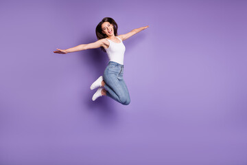 Fototapeta na wymiar Full body photo of cheerful brown haired woman jump up hands wings wear white tank-top jeans isoalted on purple color background