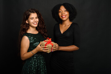 Two young women of different nationalities are smiling, holding a present for the new year and christmas on a black background. Gift exchange, giving and receiving concept