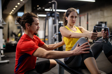 Fototapeta premium Young Hispanic personal trainer working with young woman who is exercising on a rowing machine in modern indoor gym. Healthy and active sports lifestyle
