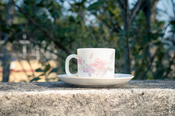 Coffee cup in morning sunlight. Summer fresh cool look. White coffee cup on saucer for hot drink on roof beam of a residential building with bokeh background.
