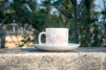 Coffee cup in morning sunlight. Summer fresh cool look. White coffee cup on saucer for hot drink on roof beam of a residential building with bokeh background.